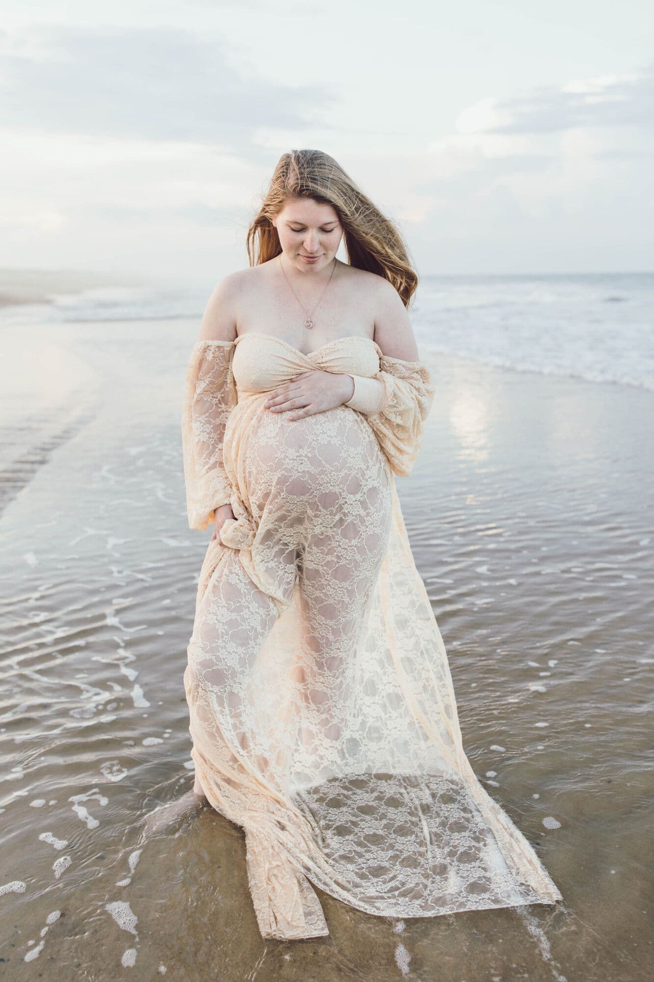Pregnant woman in lace gown standing in ocean waves during maternity session